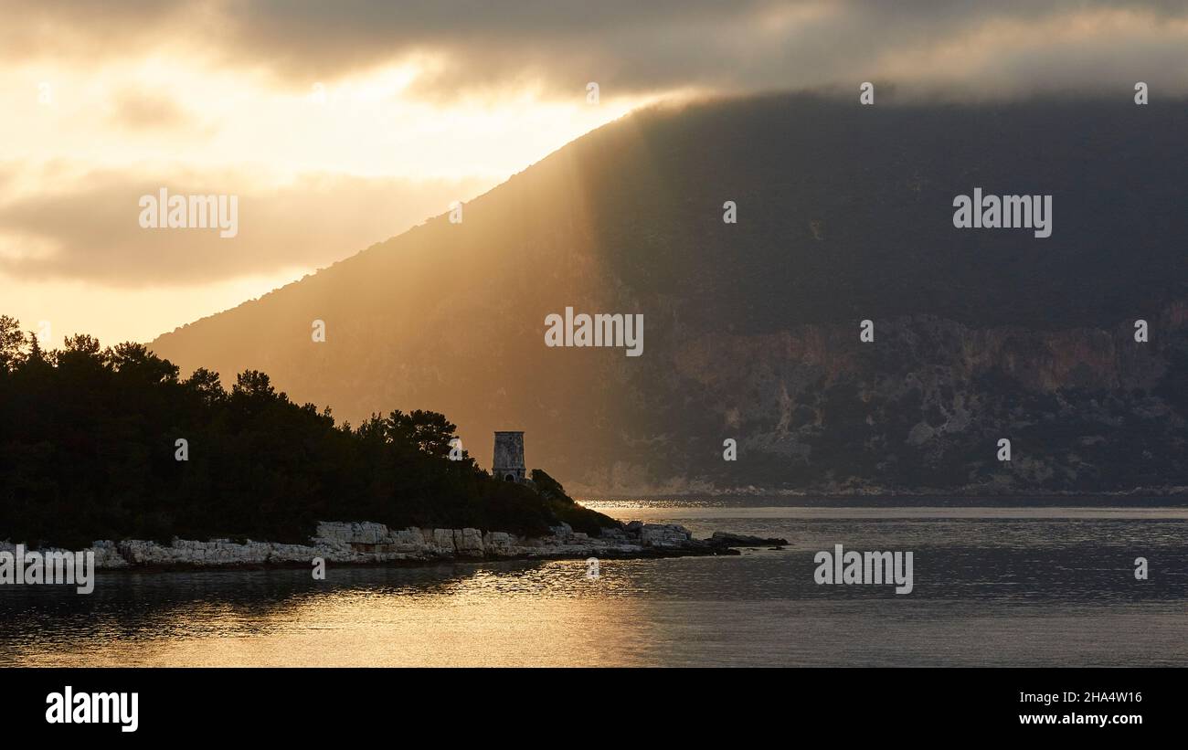 griechenland, griechische Inseln, ionische Inseln, kefalonia, fiskardo, Morgenstimmung, teilweise bewölktem Himmel, goldene Sonnenstrahlen brechen durch eine graue Wolkendecke und fallen auf die östliche Landzunge mit dem venezianischen Leuchtturm, Sonnenlicht auf dem Wasser, die ithaca-Hügel im Hintergrund Stockfoto