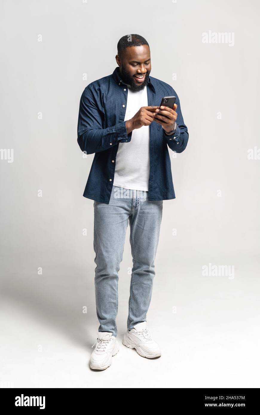 Fröhlicher und unbeschwerter afroamerikanischer Mann mit Smartphone isoliert auf grau, schwarzer Kerl verbringt Freizeit in sozialen Netzwerken, online chatten, einkaufen, mit neuen mobilen App, volle Länge, vertikal Stockfoto