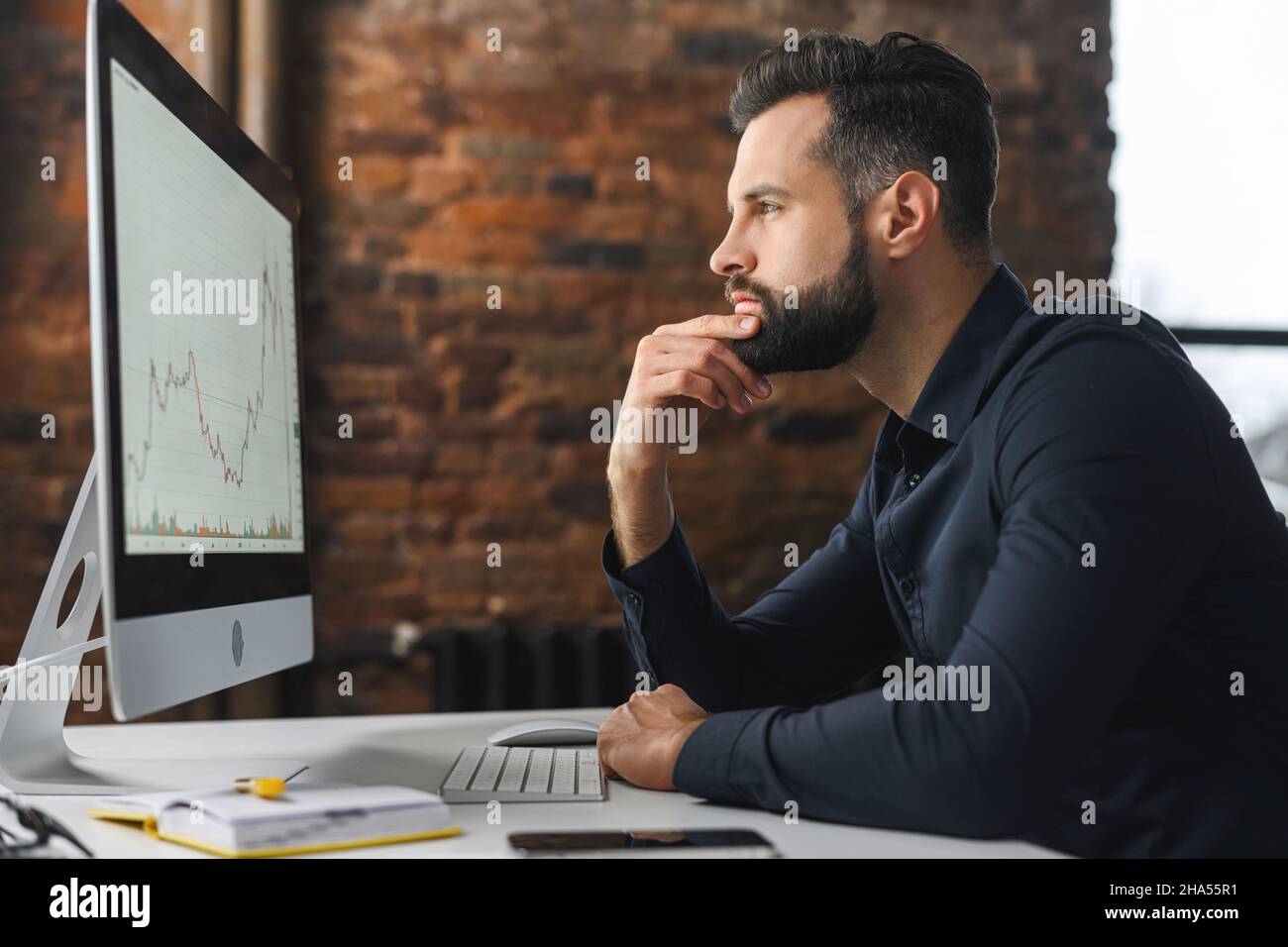 Fokussierter und besorgter Mann in elegantem Casual-Hemd, der auf den Laptop-Bildschirm blickt. Ernsthafter kaukasischer Typ, der am Schreibtisch sitzt und Zweifel spürt, während er schwierige Aufgaben löst Stockfoto