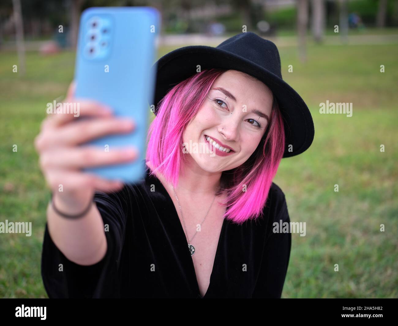 Frau mit schwarzem Hut und rosafarbenem Haar beim Selfie im Park Stockfoto