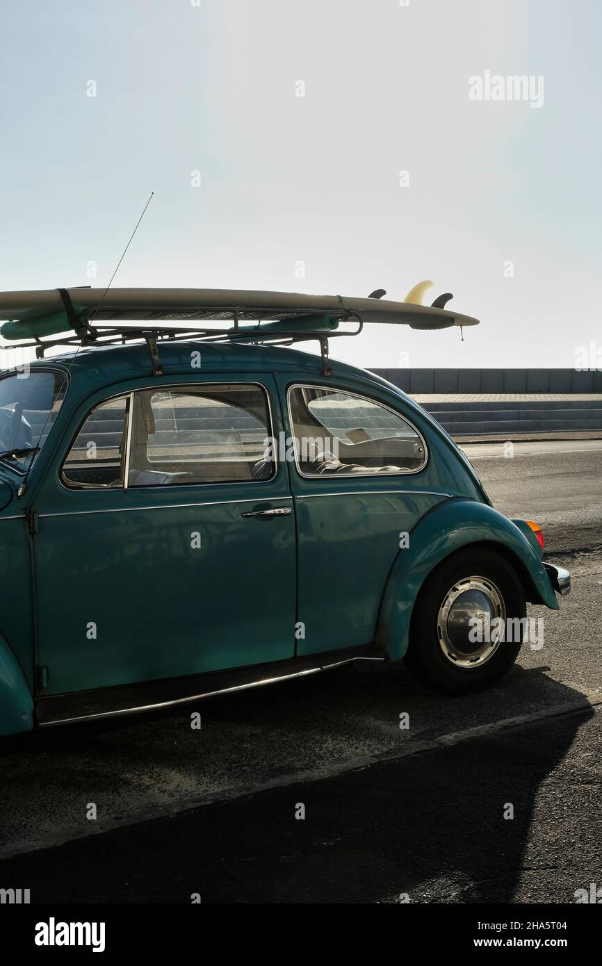 Ein alter alter alter alter Volkswagenkäfer mit einem Surfbrett auf Dachträgern am Straßenrand. Am späten Nachmittag mit Strandatmosphäre und Kopierbereich. Stockfoto