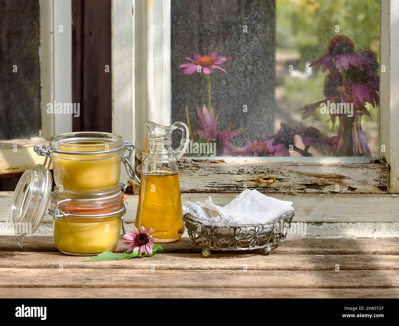 Zwei mit Echinacea-Salbe gefüllte Vorratsgläser und eine Glaskaraffe aus Pflanzenöl stehen zusammen mit einem Metallkorb und textilen Taschentüchern auf einem Holztisch vor einem Werkstattfenster Stockfoto