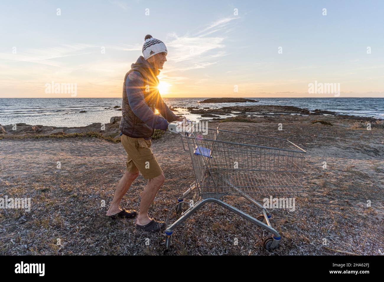 Wie viel Konsum kann die Natur vertragen? Mann mit Einkaufswagen am Meer Stockfoto