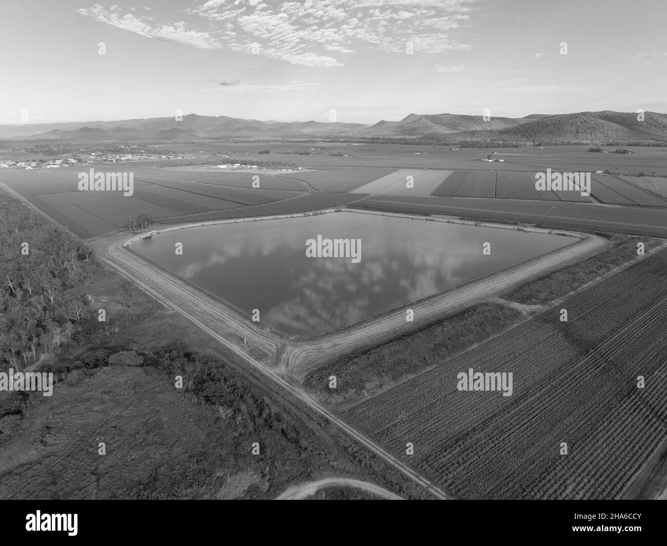 Monotone eines quadratischen ländlichen Staudamms zwischen Zuckerrohrfeldern mit einem Township in der Ferne Stockfoto