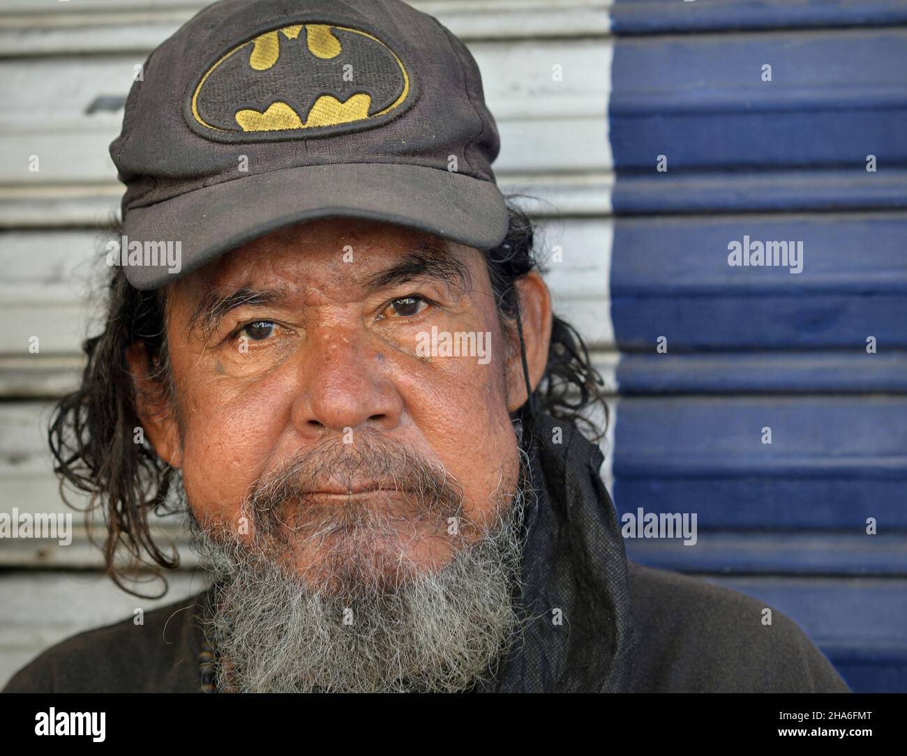 Älterer, robuster, obdachloser mexikanischer Mann mit grauem Bart und zerzaust behaartem Haar trägt eine abgenutzte Baseballmütze und blickt auf den Betrachter. Stockfoto