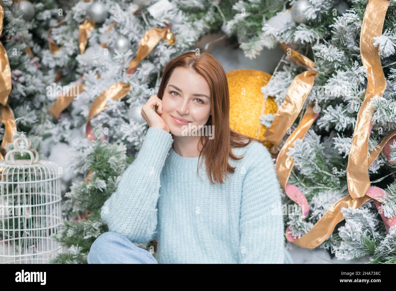 Neujahrs-Porträt der schönen lächelnden kaukasischen jungen Frau in gemütlichen Wolle warm hellblau Pullover. Weihnachtsbaum im Hintergrund. Stockfoto