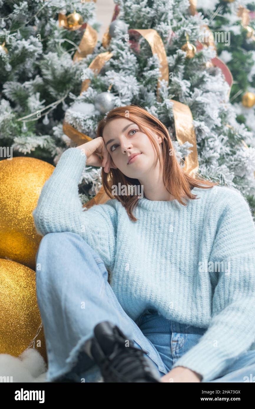 Neujahrsporträt der schönen träumenden kaukasischen jungen Frau in kuscheligen Wolle warmen hellblauen Pullover. Weihnachtsbaum im Hintergrund. Stockfoto