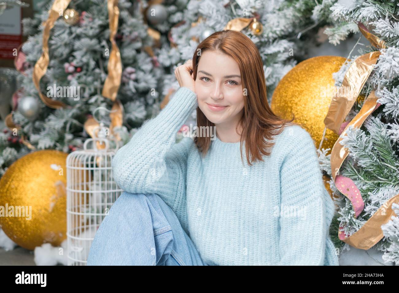 Neujahrs-Porträt der schönen lächelnden kaukasischen jungen Frau in gemütlichen Wolle warm hellblau Pullover. Weihnachtsbaum im Hintergrund. Stockfoto