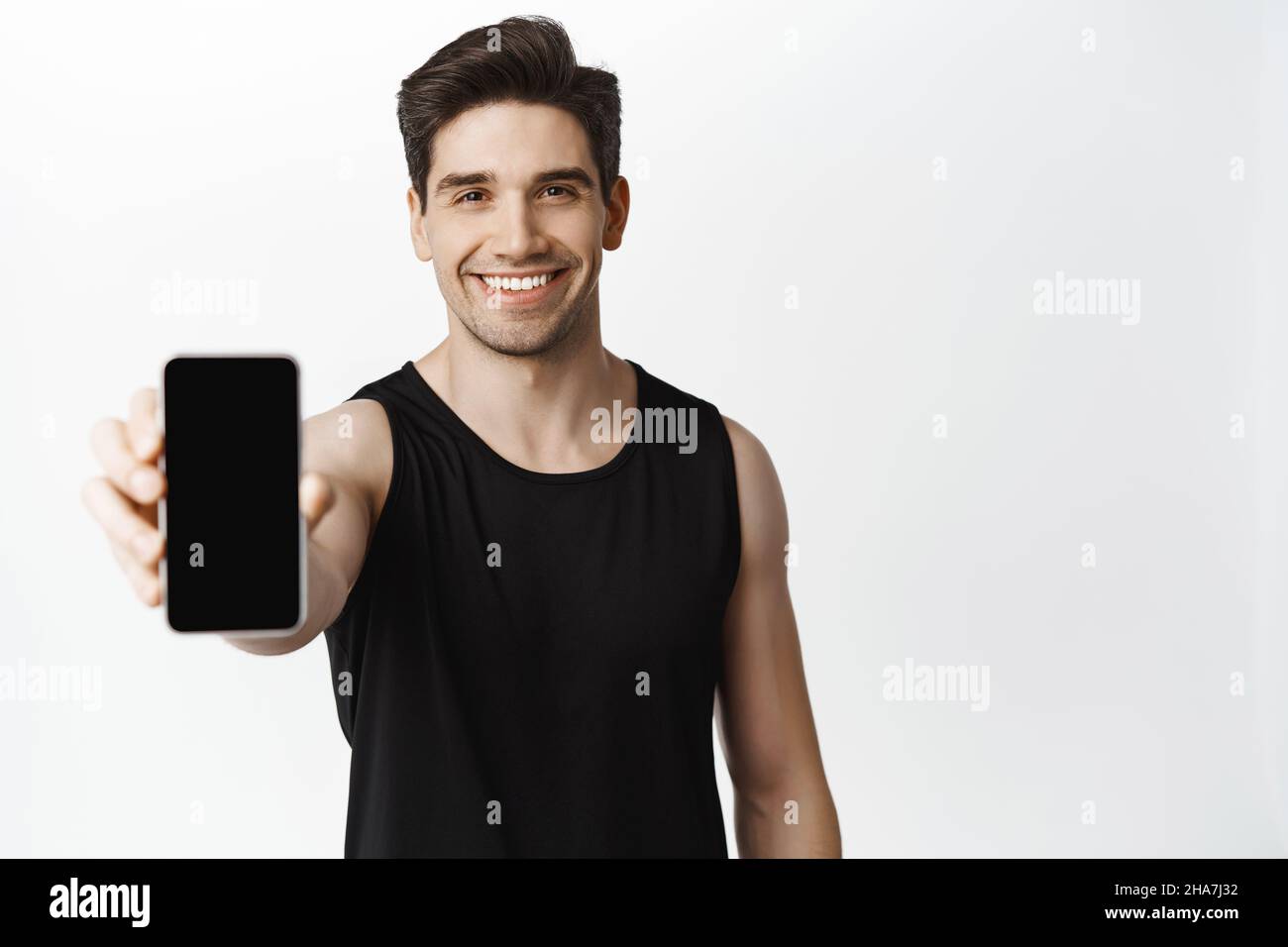 Schöner starker Kerl zeigt Smartphone-Bildschirm und lächelt. Männlicher Athlet demonstriert Workout-App auf Mobiltelefon, weißer Hintergrund Stockfoto
