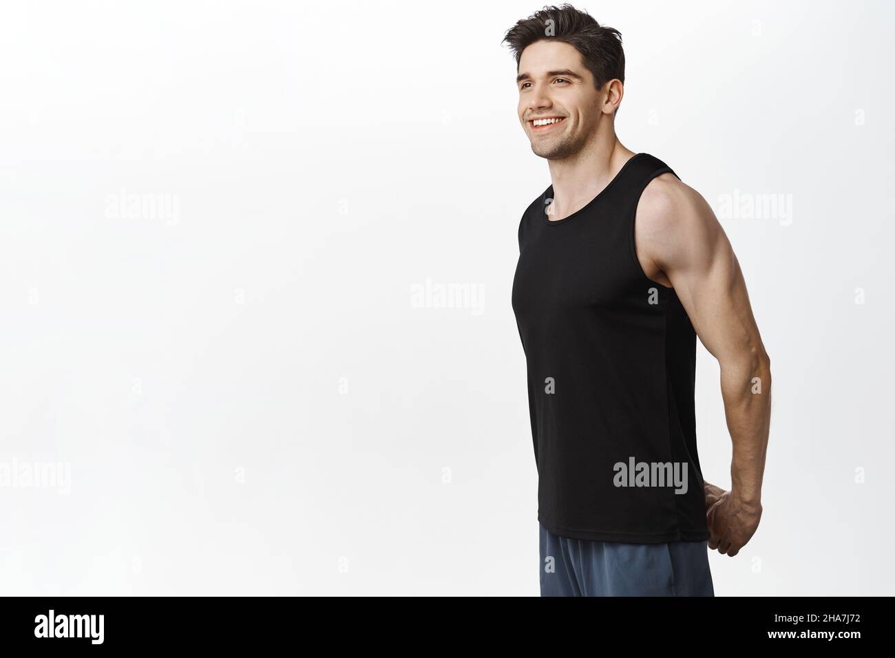 Lächelnder männlicher Athlet, Fitness-Typ, der die Arme hinter dem Rücken streckt, Aufwärmen für Trainingseinheiten, Fitnessübungen, weißer Hintergrund Stockfoto