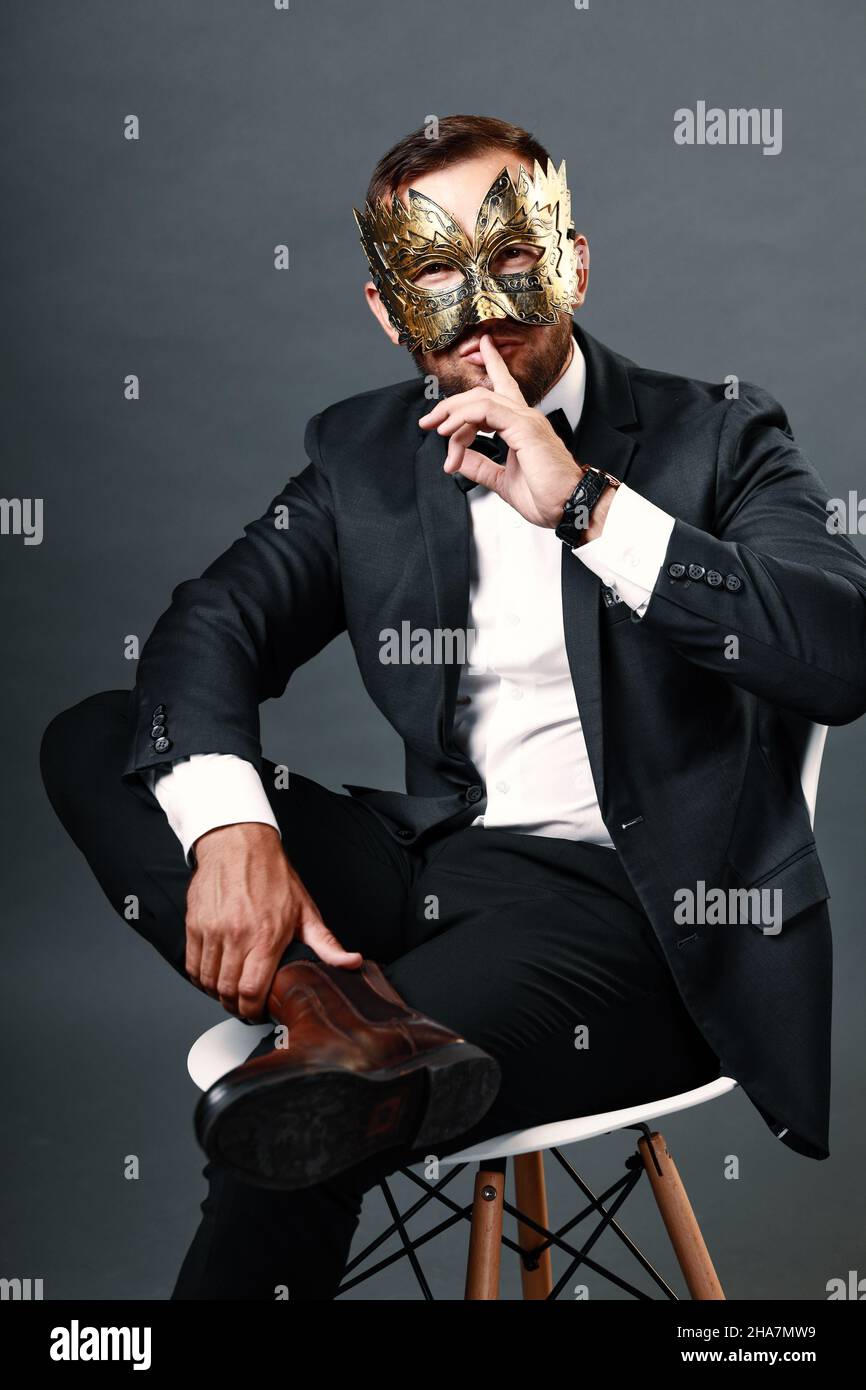 Der Mann im Business-Anzug sitzt auf dem Stuhl und trägt eine Karnevalsmaske auf seinem Gesicht. Studio-Porträt auf grauem Hintergrund. Ein junger Mann von 25-30 Jahren versteckt sein Gesicht unter Maske. Listiger und hinterlistiger Geschäftsmann. Trickster. Stockfoto