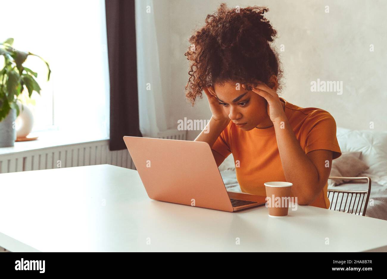 Müde flustrierte afrikanisch abstammende junge Mädchen, die vor dem Laptop am Schreibtisch sitzt, während sie irritierend auf den Computerbildschirm mit den Händen schaut, die ihren Kopf halten Stockfoto