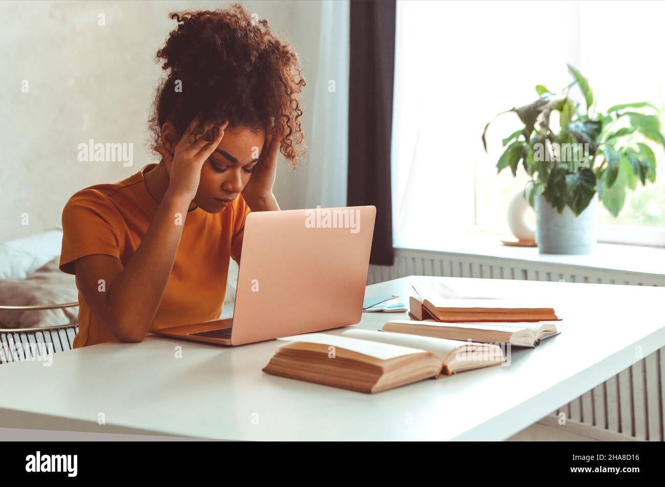 Müde flustrierte afrikanisch abstammende junge Mädchen, die vor dem Laptop am Schreibtisch sitzt, während sie irritierend auf den Computerbildschirm mit den Händen schaut, die ihren Kopf halten Stockfoto