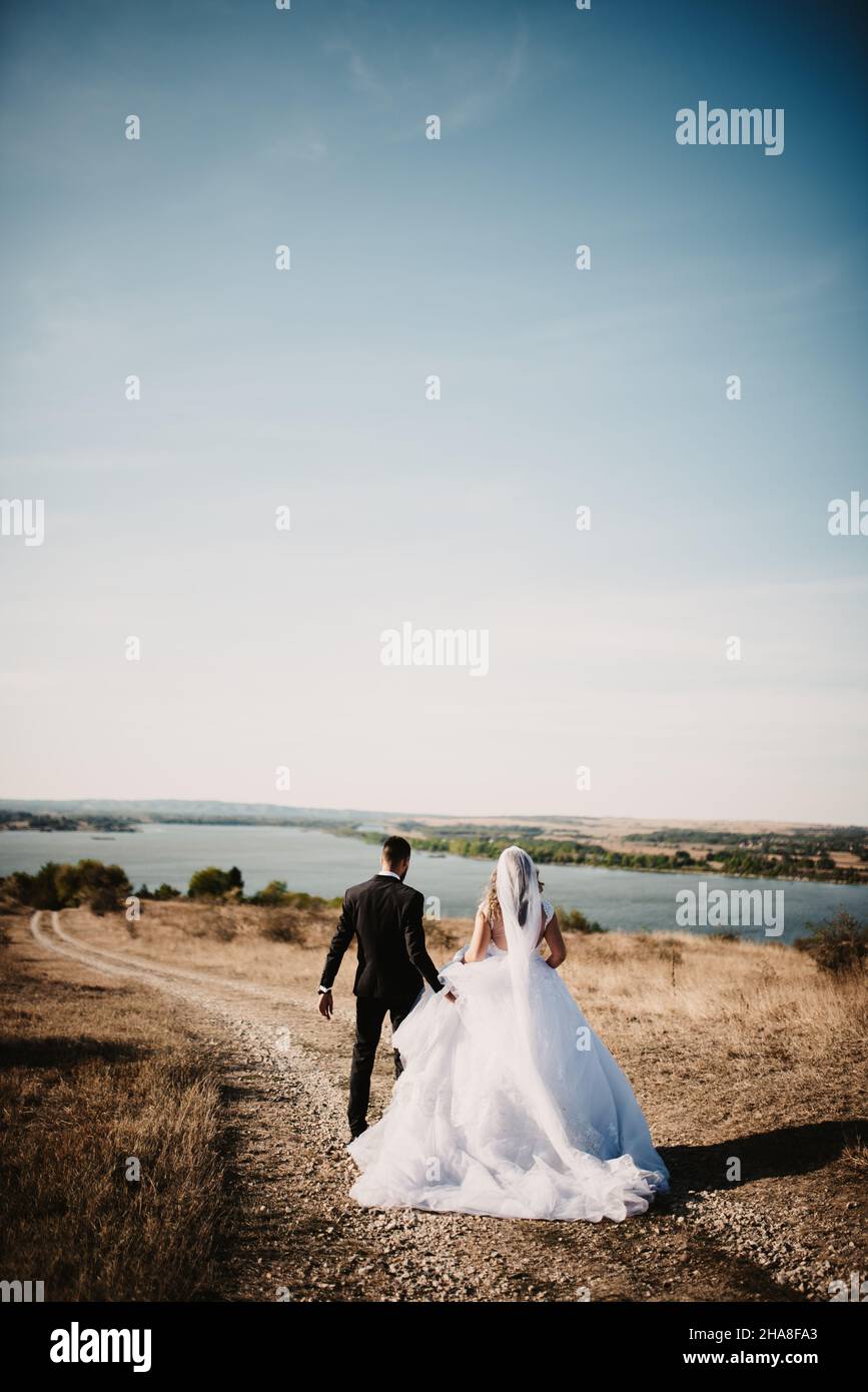 Ruhige Aussicht auf eine Braut in einem schönen Hochzeitskleid und den Bräutigam, der am Ufer eines Sees spazieren geht Stockfoto