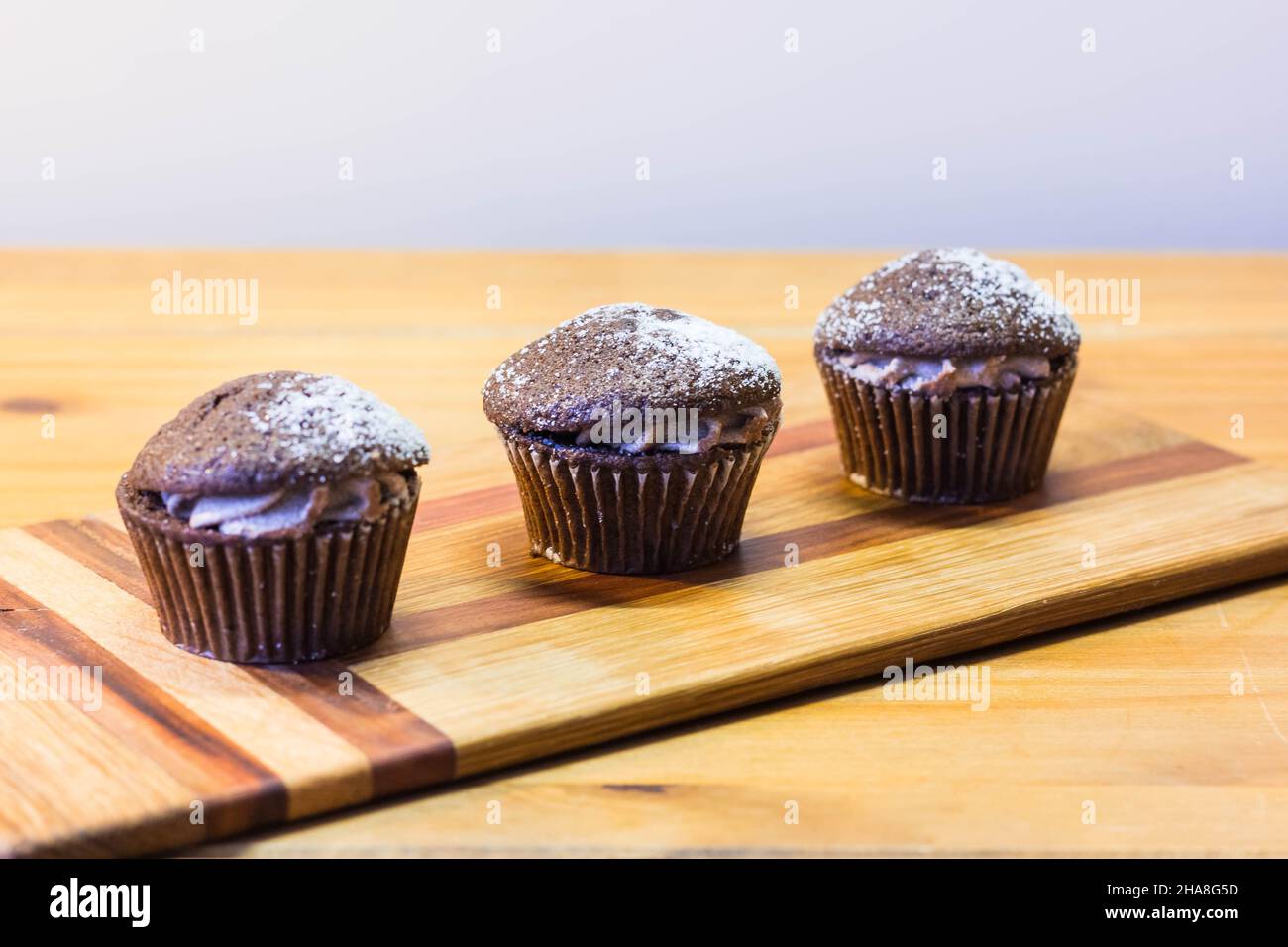 Drei Schokoladen-Victoria-Sponge-Mini-Cup-Kuchen mit Kakaocreme gefüllt und mit Zucker auf Kirschholzbrett bestäubt Stockfoto