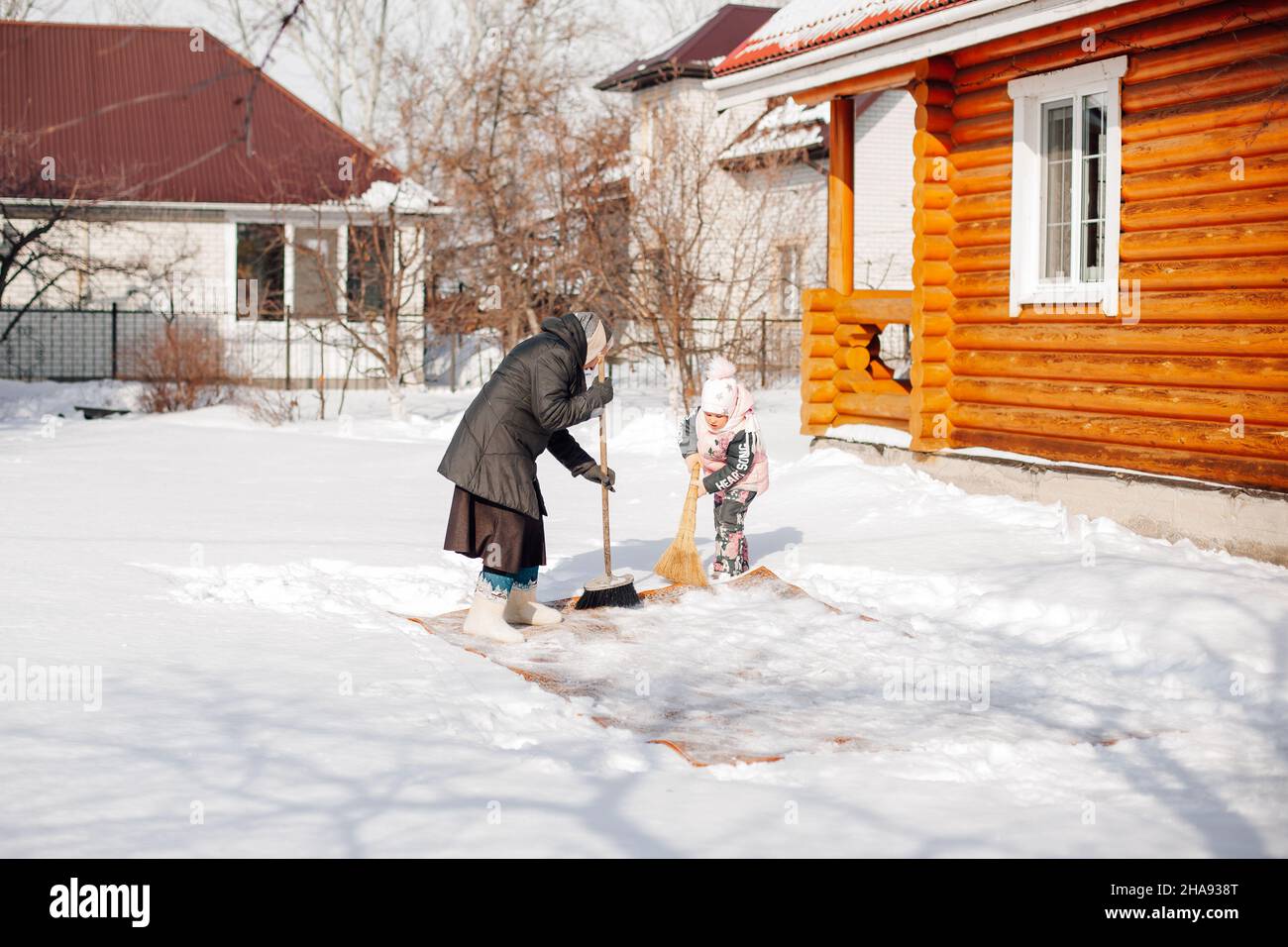 Frau und Kind fegen Schnee mit Besen weg. Die Familie beschäftigt sich mit der ökologischen Teppichreinigung nach traditioneller Methode mit Hilfe von Neuschnee in der Nähe der Hütte Stockfoto