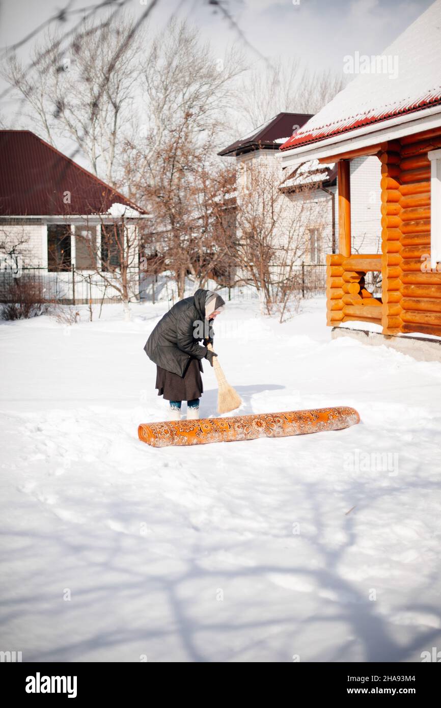 Kaukasische Frau reinigt Teppich. Ältere Frau in Schal und Filzstiefeln fegt Schnee vom Teppich im Hinterhof in der Nähe eines Holzhauses, in dem sie verlobt ist Stockfoto