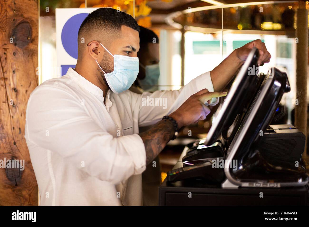 Junger lateinamerikanischer Restaurantmitarbeiter mit medizinischer Maske, der die Rechnung an der Kasse vorbereitet. Stockfoto