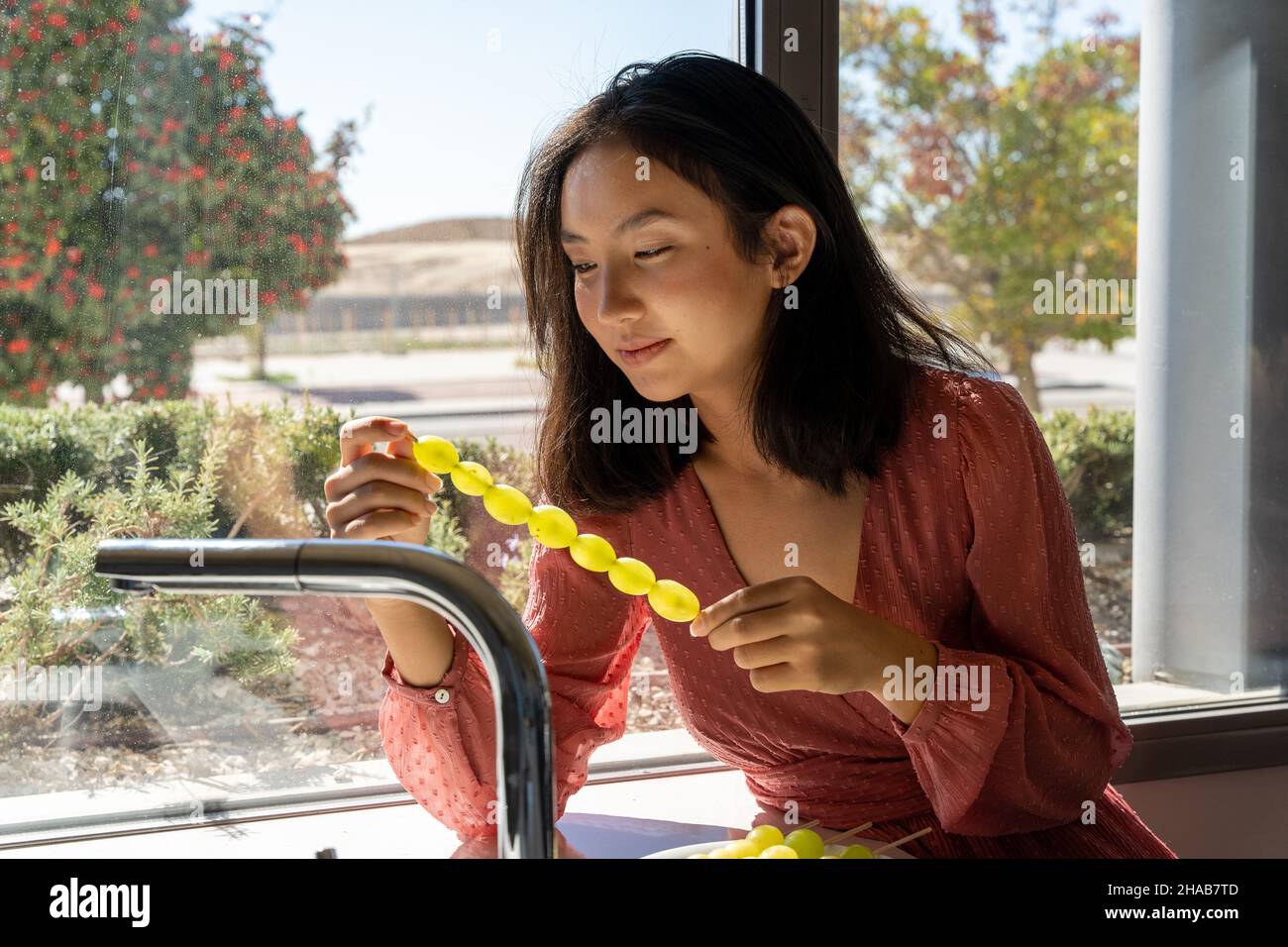 Frau, die auf den Spieß der Trauben schaut, die sie vorbereitet, horizontales Bild von der Taille nach oben, beleuchtet durch Sonnenlicht von außen Stockfoto