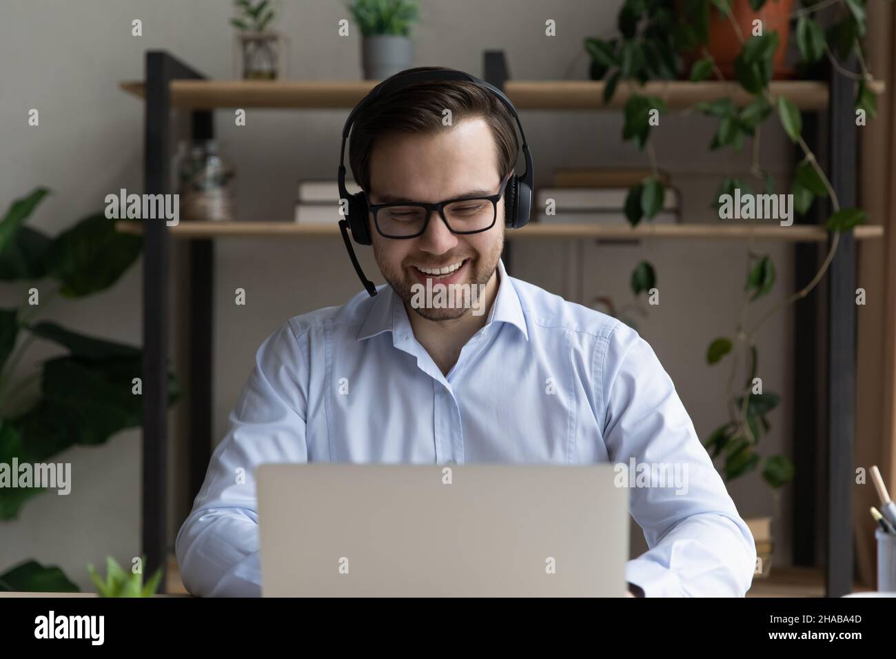 Ein fröhlicher Geschäftsmann im Headset sitzt am Schreibtisch und schaut auf den Laptop Stockfoto