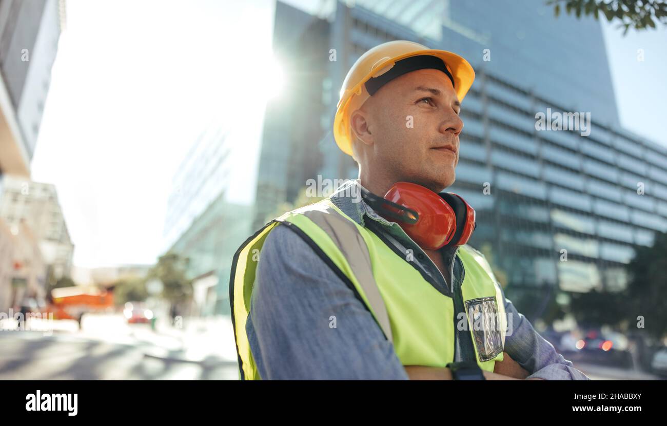 Nachdenklicher Arbeiter, der in der Stadt steht. Der Bauarbeiter mittleren Erwachsenen schaute nachdenklich weg, während er mit gekreuzten Armen im Freien stand. Blau c Stockfoto