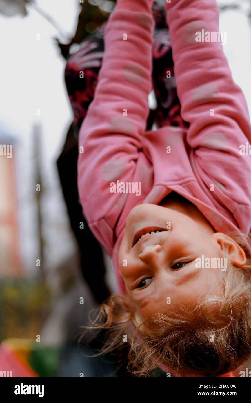 Baby 4 Jahre alt im Freien auf dem Spielplatz. Das Kind zieht sich an den horizontalen Balken nach oben. Das Mädchen hängt auf dem Kopf. Stockfoto