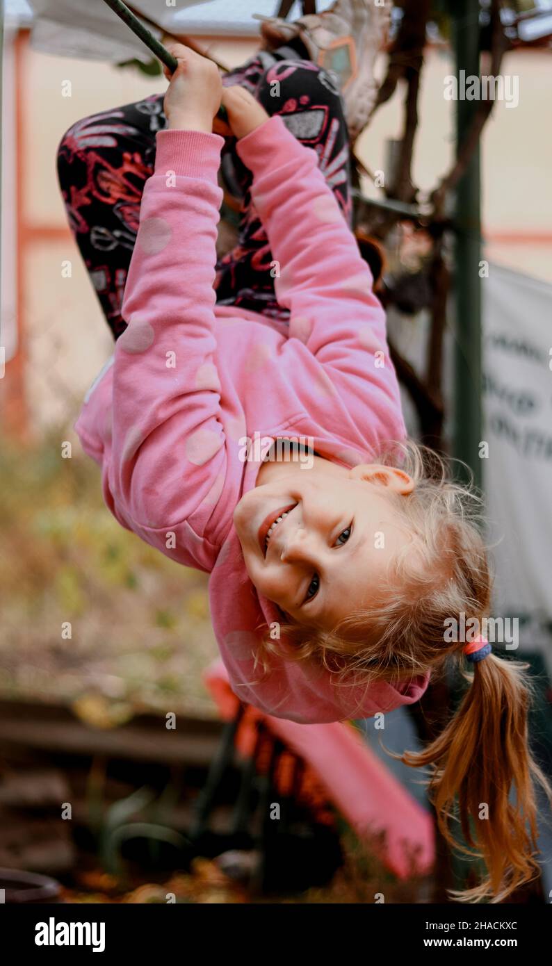 Baby 4 Jahre alt im Freien auf dem Spielplatz. Das Kind zieht sich an den horizontalen Balken nach oben. Das Mädchen hängt auf dem Kopf. Stockfoto