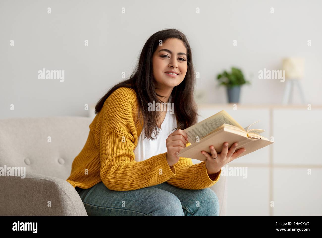 Freie Zeit zum Lesen. Glückliche arabische Frau, die in einem bequemen Sessel mit Buch sitzt, sich am Wochenende zu Hause ausruht, freier Platz Stockfoto