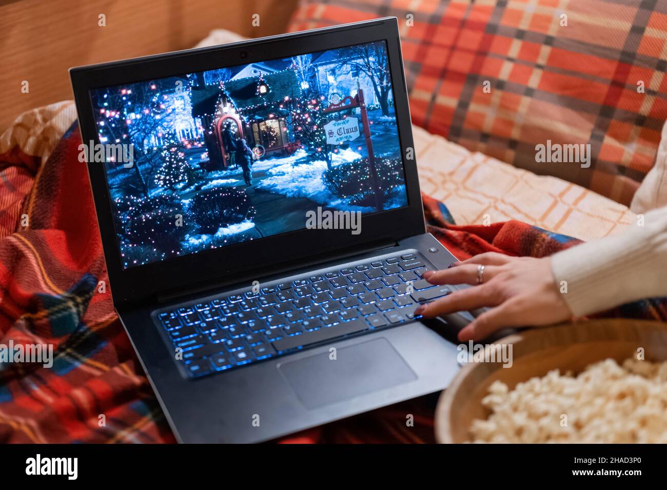 Laptop-Display mit einem festlichen Weihnachtsfilm „Home Alone“ auf einem roten karierten karierten karierten karierten Bett zu Hause und einem Video zum Zurückspulen der weiblichen Hand Stockfoto