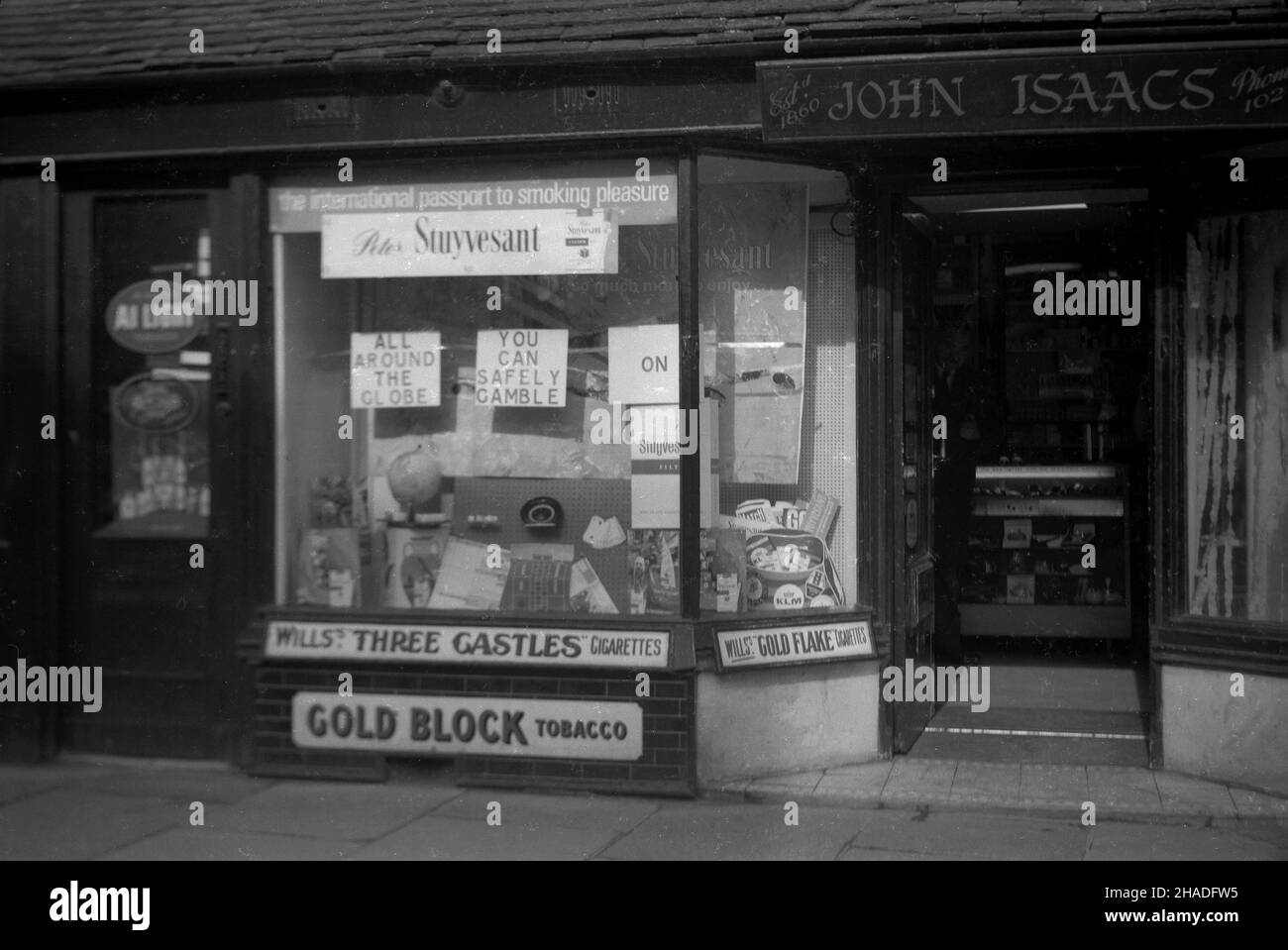 1958, Außenansicht des Schaufensters eines Tabakwarenhandels, John Isaacs, Half Moon Street, Sherborne, Dorset, England, Großbritannien. John Isaacs war ursprünglich Friseur und die Räumlichkeiten zu einer Zeit, kombiniert mit dem Verkauf von Tabak. Im Fenster, Werbung für Peter Stuyvesant Zigaretten, "der internationale Pass zum Rauchvergnügen" und "rund um den Globus, können Sie sicher auf Peter Stuyvesant spielen." Die Marke wurde ‘in Deutschland gegründet und mit ihrer „amerikanischen Mischung“ aus Tabak aus verschiedenen Ländern als international propagiert. Gezeigt wurden auch Wills Three Castles Cigarettes & Gold Block Tabak. Stockfoto 1958, Außenansicht des Schaufensters eines Tabakwarenhandels, John Isaacs, Half Moon Street, Sherborne, Dorset, England, Großbritannien. John Isaacs war ursprünglich Friseur und die Räumlichkeiten zu einer Zeit, kombiniert mit dem Verkauf von Tabak. Im Fenster, Werbung für Peter Stuyvesant Zigaretten, "der internationale Pass zum Rauchvergnügen" und "rund um den Globus, können Sie sicher auf Peter Stuyvesant spielen." Die Marke wurde ‘in Deutschland gegründet und mit ihrer „amerikanischen Mischung“ aus Tabak aus verschiedenen Ländern als international propagiert. Gezeigt wurden auch Wills Three Castles Cigarettes & Gold Block Tabak. Stockfoto