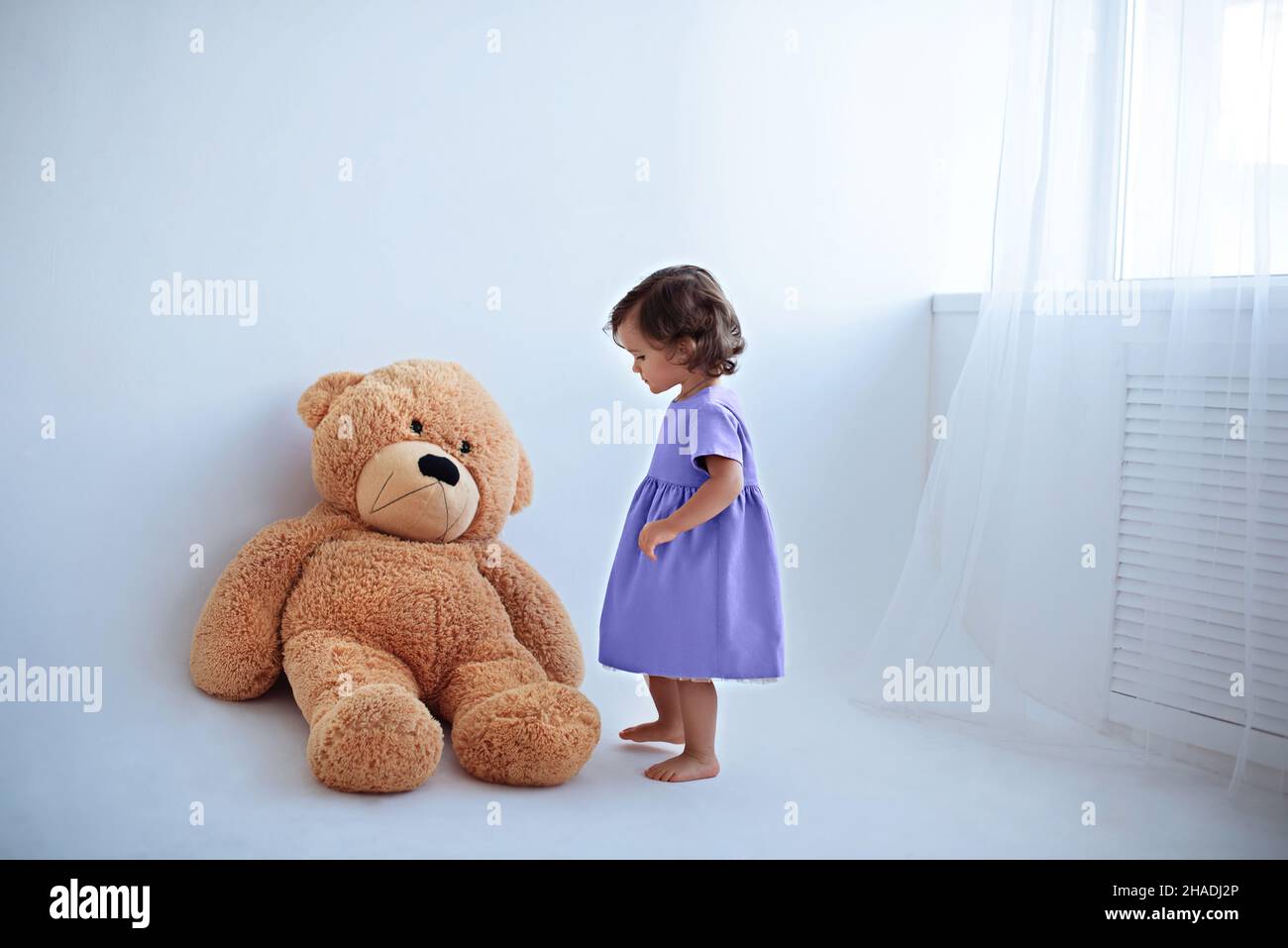 Kleines Mädchen im lila Lavendelkleid steht neben einem großen Teddybären Stockfoto