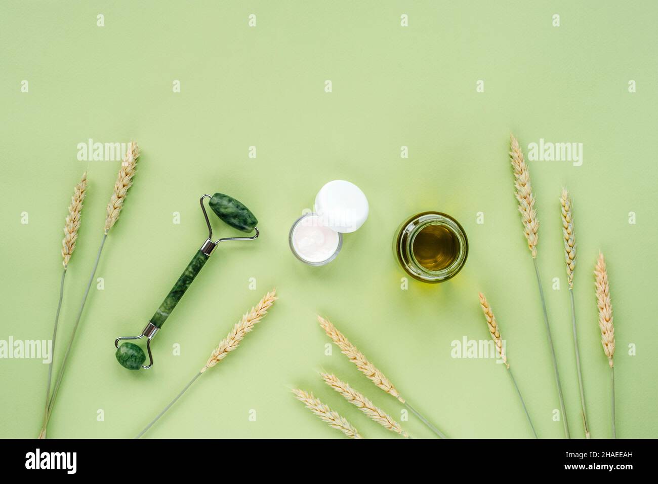 Kosmetisches Öl in Glasflasche, Cremeglas, Gesichtswalze und Ähren aus Weizen auf hellgrünem Hintergrund. Hautpflege, natürliche Bio-Kosmetik-Konzept. Top Vie Stockfoto