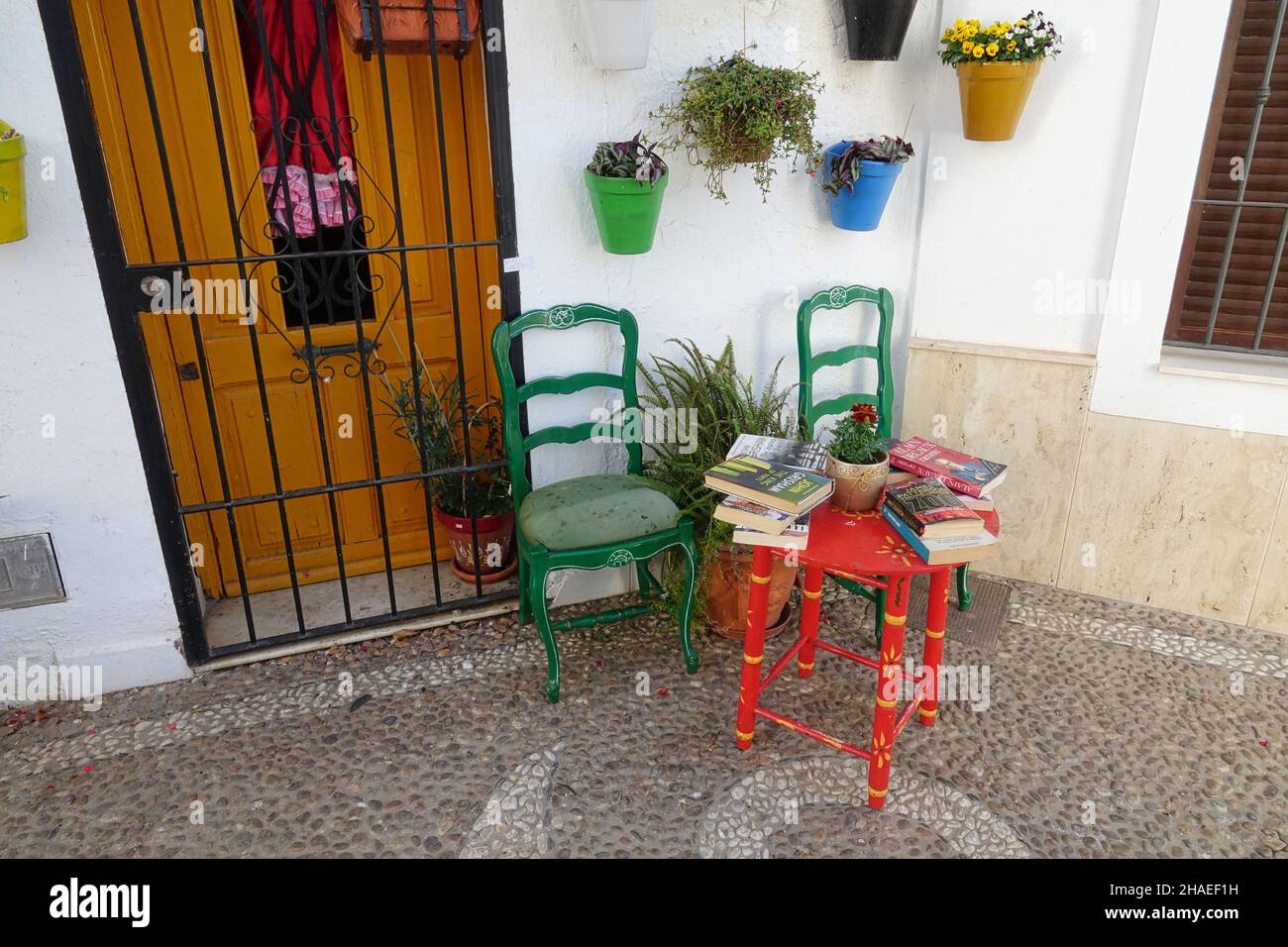 Nerja, Spanien: Gebrauchte Bücher auf einem roten Tisch neben zwei grünen Stühlen an der Tür eines typisch andalusischen Hauses mit einer weißen Fassade mit Blumentöpfen Stockfoto