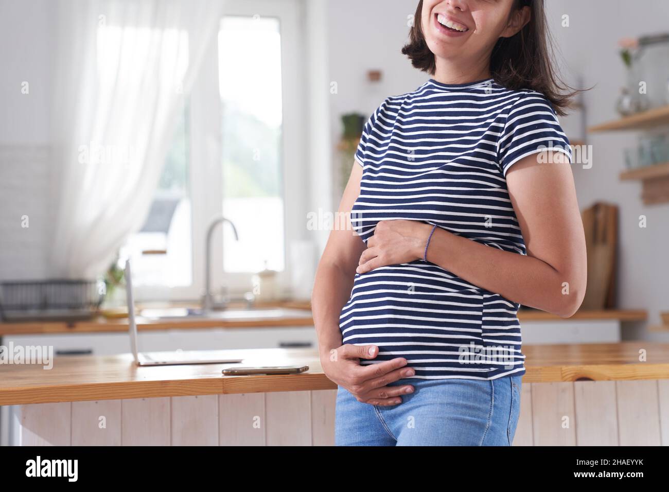 Eine schwanger Frau wickelt ihre Arme um ihren Bauch. Eine Frau im dritten Trimester der Schwangerschaft steht in der Küche. Das Konzept des Wartens auf ein Kind. Hochwertige Fotos Stockfoto