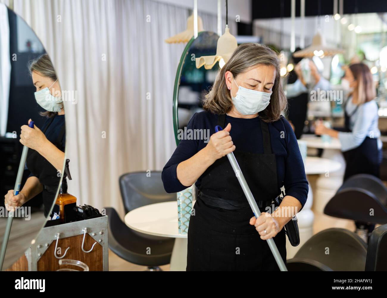 Arbeitsprozess im Schönheitssalon während der Pandemie-Situation. Ältere weibliche Friseurin in Schutzmaske Reinigung Arbeitsbereich, fegenden Boden Stockfoto