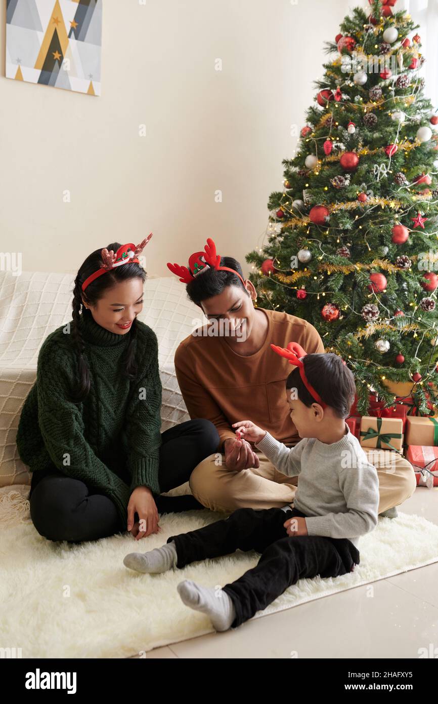 Junge Eltern spielen mit dem kleinen Sohn, wenn sie zu Hause neben dem Weihnachtsbaum auf dem Boden sitzen Stockfoto