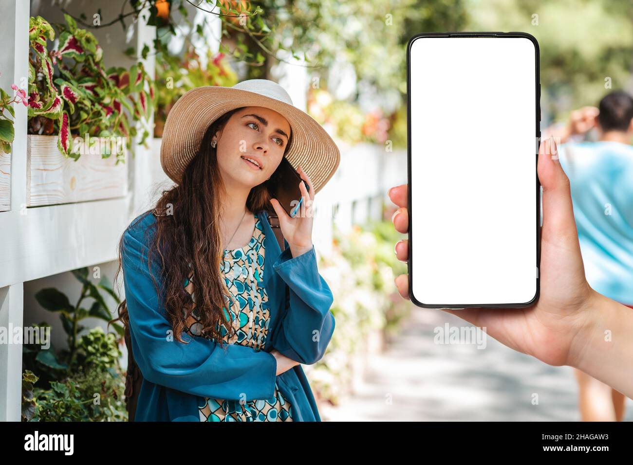 Eine Frau mit Strohhut telefoniert. Im Hintergrund eine Stadtstraße. Im Vordergrund und Hand hält ein Smartphone, Mock up. Kommunikation Stockfoto