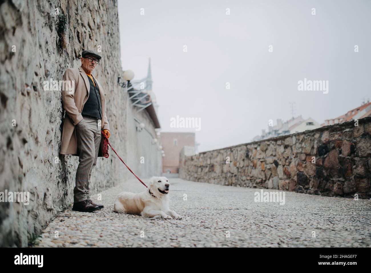 Älterer Mann, der auf die Kamera schaute und sich an die Wand lehnte, als er seinen Hund in der Stadt draußen spazieren ging. Stockfoto