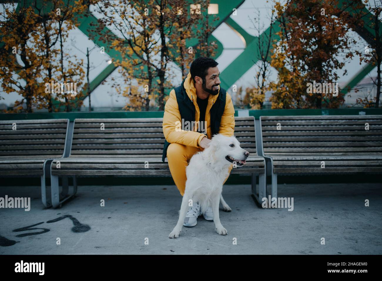 Glücklicher junger Mann, der im Herbst auf der Bank sitzt und seinen Hund im Freien in der Stadt hält. Stockfoto
