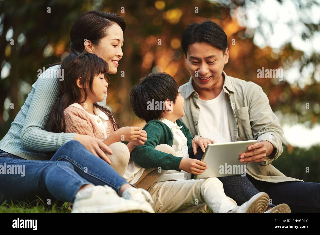 Glückliche asiatische Familie mit zwei Kindern, die eine gute Zeit auf Gras sitzen und sich im Stadtpark bei Sonnenuntergang entspannen Stockfoto