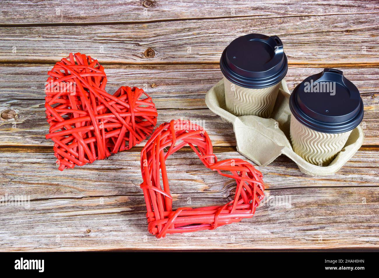 Auf einem hölzernen Hintergrund befinden sich rechts zwei Tassen Kaffee und links zwei große dekorative gewebte Herzen in verschiedenen Formen für den h Stockfoto