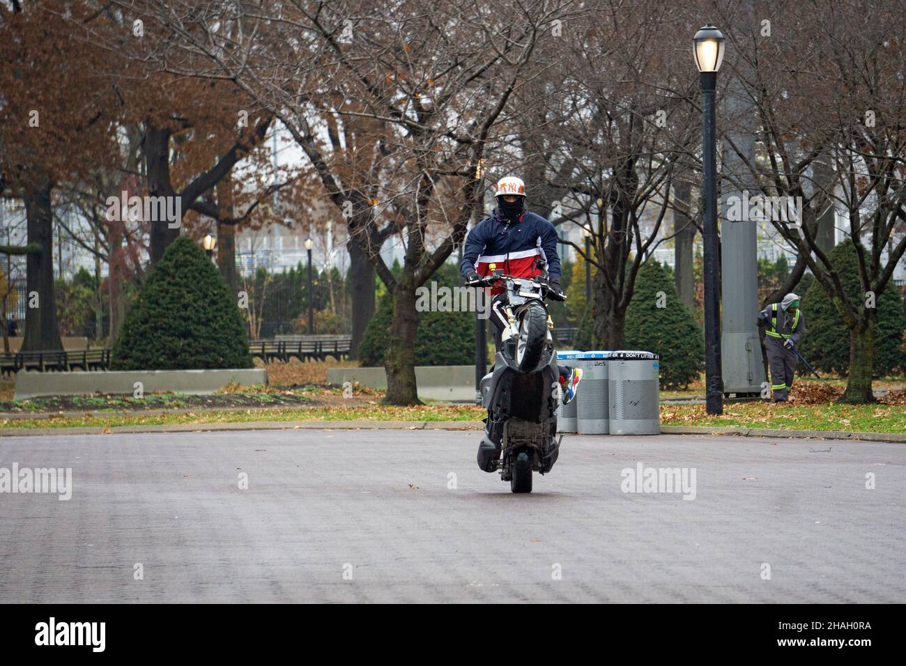 Ein Teenager-Biker mit Helm und Maske macht ein Wheelie auf seiner Yamaha TMax, während er auf dem Sitz steht. In einem Park in Queens, New York City. Stockfoto