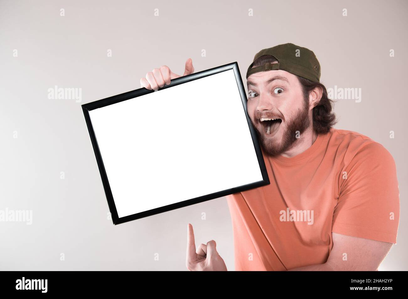Junger bärtiger Mann, der ein lustiges Gesicht macht und ein leeres Schild hält. Studio auf grauem Hintergrund aufgenommen. Stockfoto
