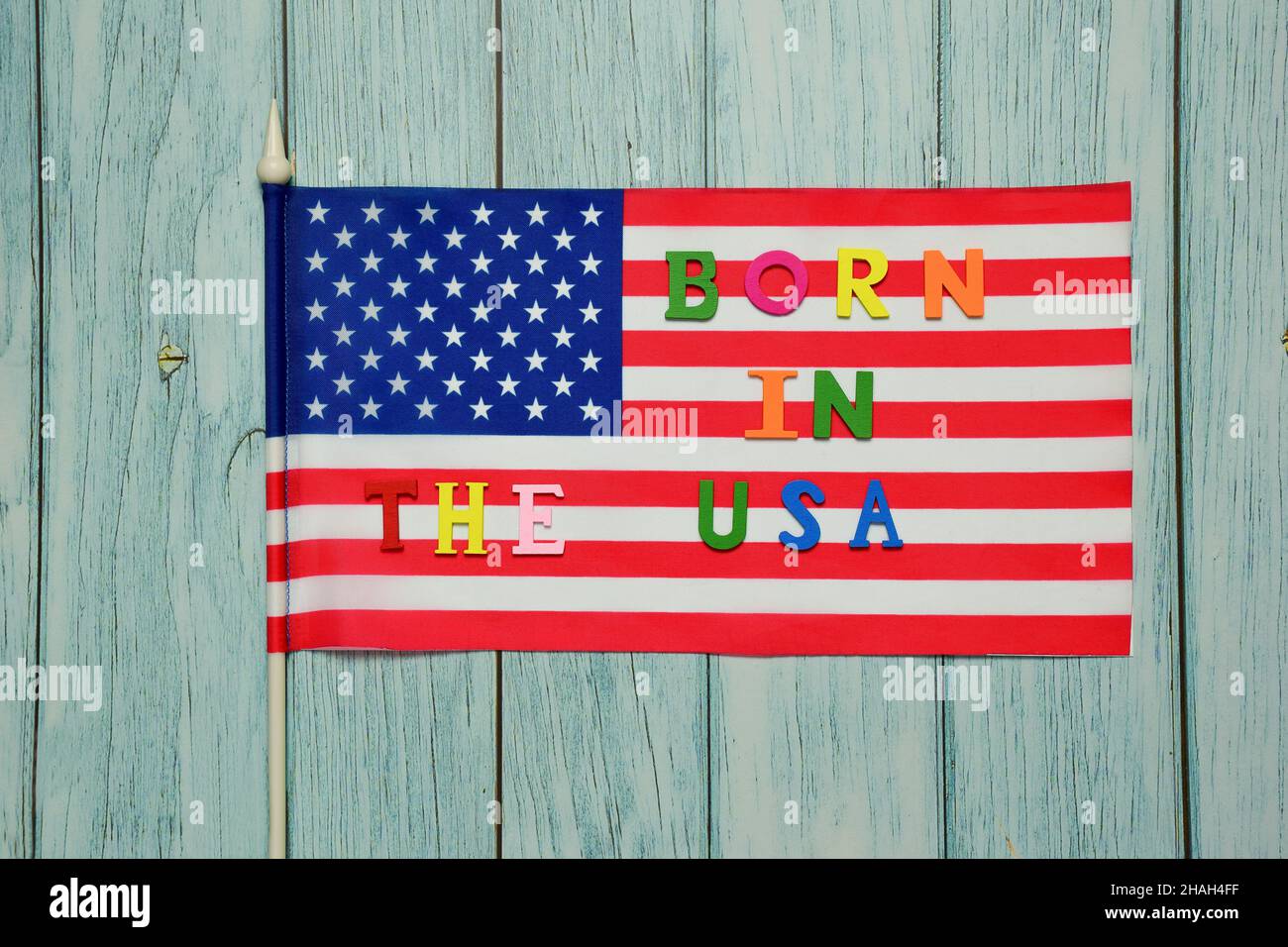 Auf der Flagge der USA ist der Text BORN IN THE USA mit bunten Buchstaben vor dem Hintergrund eines Holzbohlenhintergrunds gesäumt Stockfoto