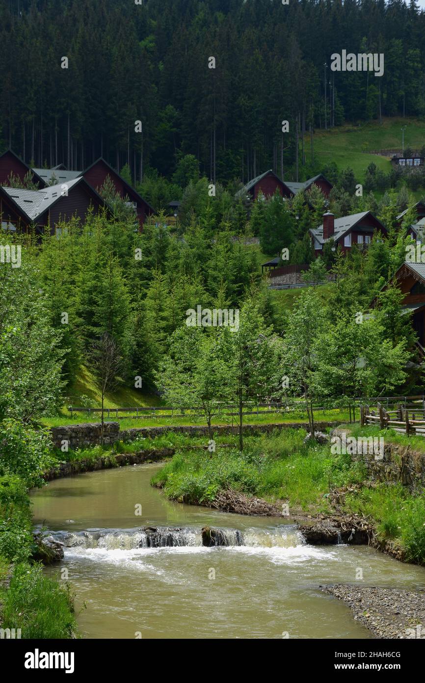 Im Vordergrund fließt ein Fluss mit schlammigem Wasser und einem kleinen Wasserfall. Dahinter sehen Sie die Hotelhäuser in der Nähe des Berges hinter grünen Bäumen Stockfoto