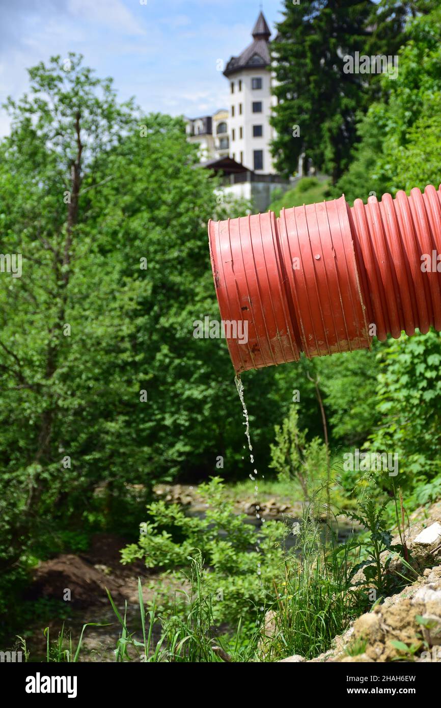 Aus einem roten Kunststoff-Abwasserrohr fließt ein dünner Wasserstrom vor einem verschwommenen Hintergrund aus Bäumen und einem Gebäude Stockfoto
