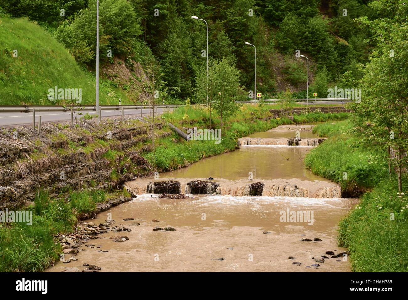 Schmutziger, brauner Wasserstrom in einem Fluss mit kleinen Wasserfällen in der Nähe der Straße aufgrund von Überschwemmungen Stockfoto