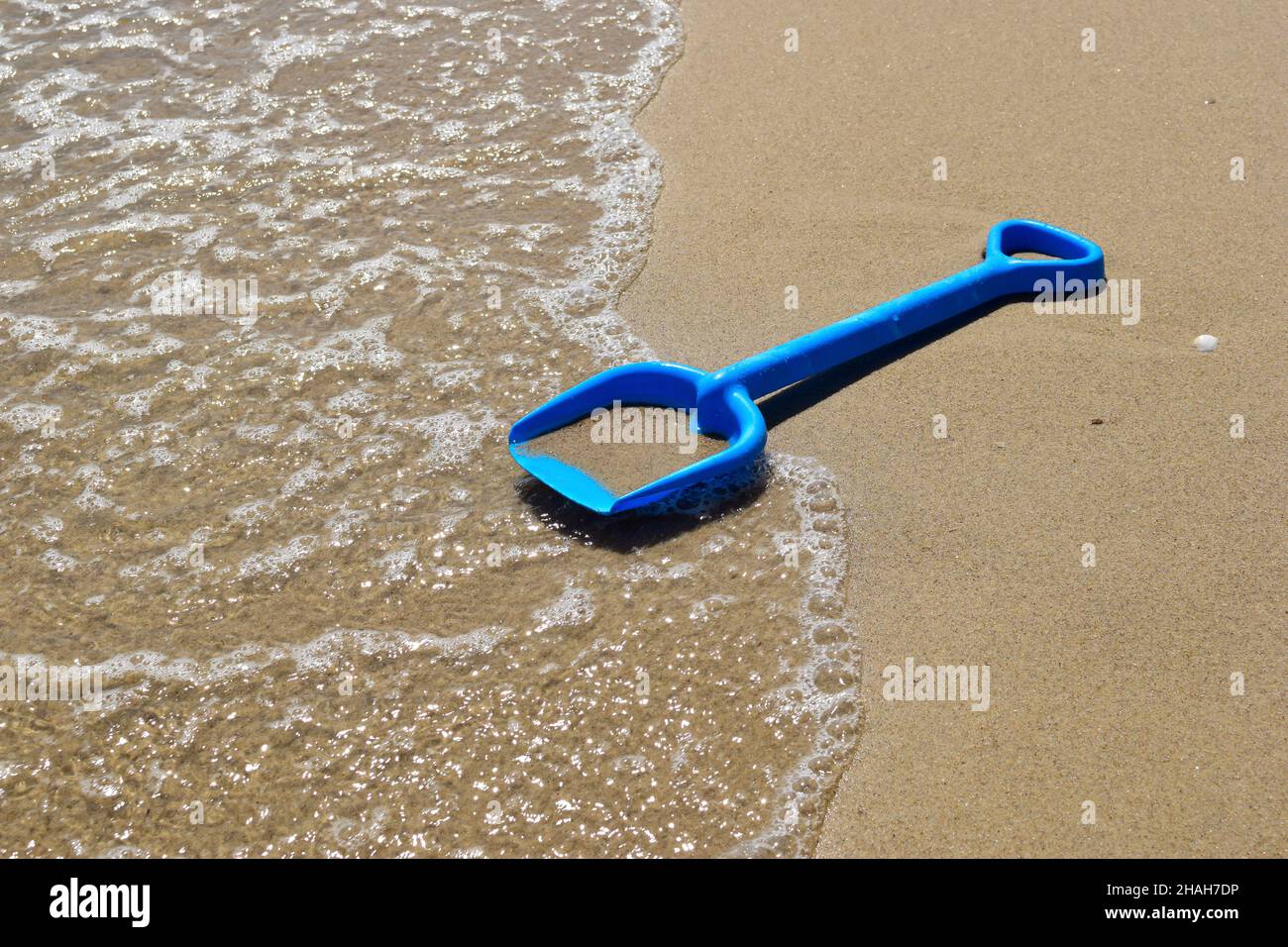 Am Sandstrand des Meeres liegt eine Kinderschaufel vor dem Hintergrund einer schäumenden Welle. Nahaufnahme Stockfoto