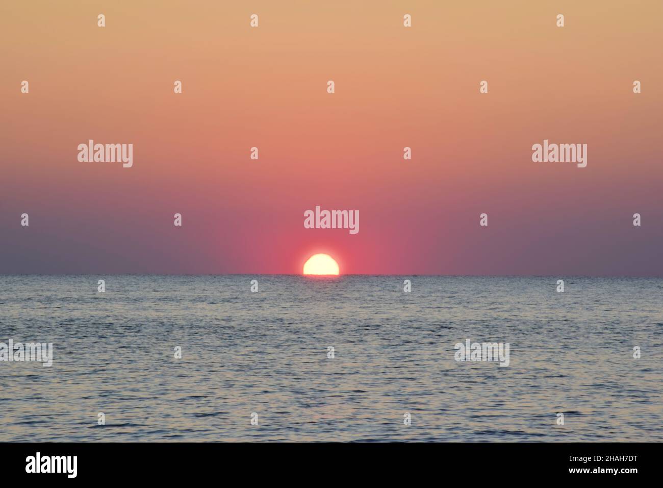 Die Morgensonne stieg aus dem ruhigen Meer zur Hälfte auf. Vor dem Hintergrund eines klaren rosa Himmels und kleiner Wellen Stockfoto