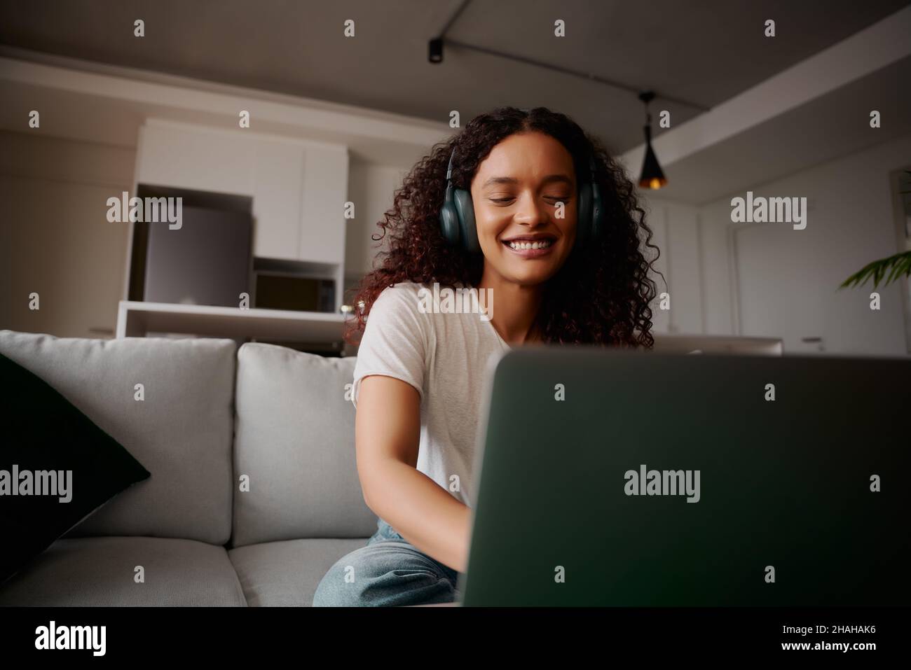 Multikulturelle Frau, die auf dem Laptop lächelt, während sie auf dem Sofa sitzt und Musik hört Stockfoto